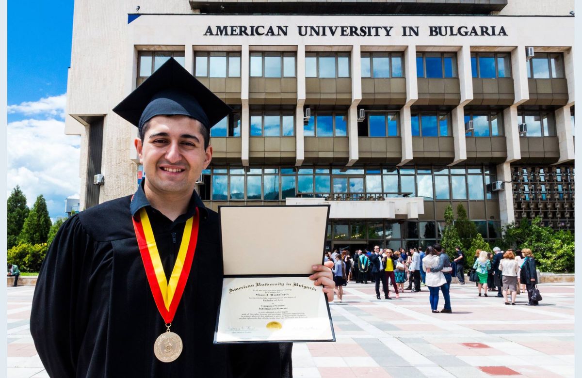 Shamil on his graduation posing in front of Main Building holding his diploma